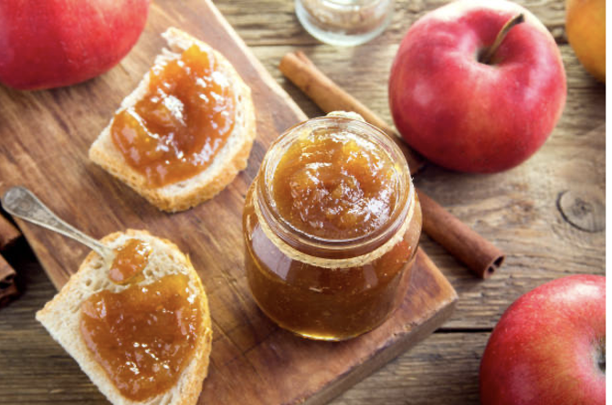 Apple butter in a jar surrounded by fresh apples and cinnamon sticks.